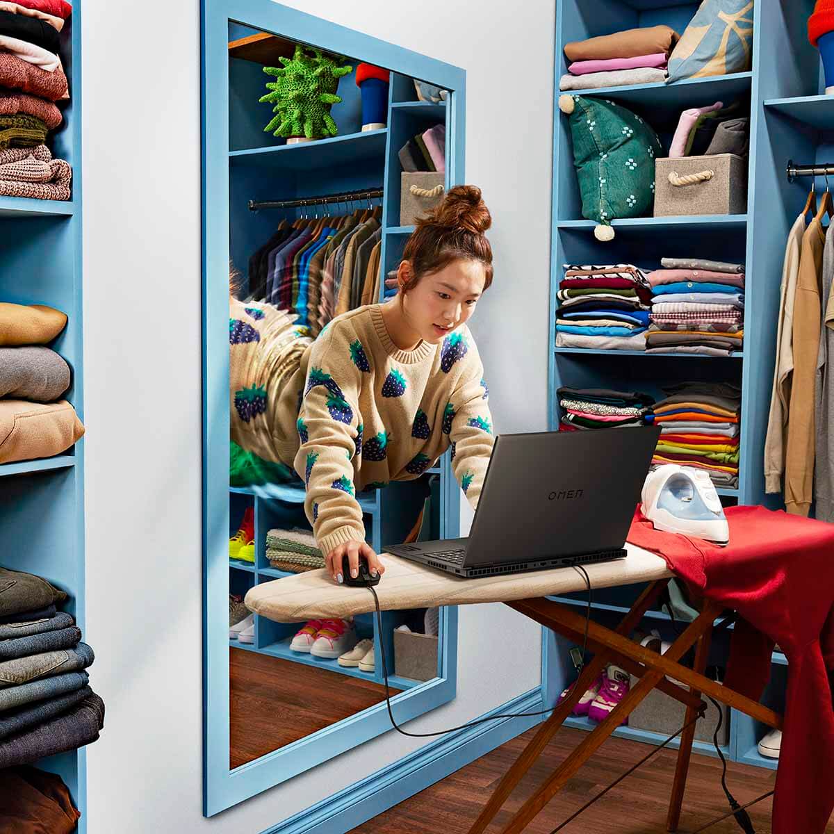 A woman leaning out of the mirror in the dressing room reaches for a laptop standing on the ironing board
